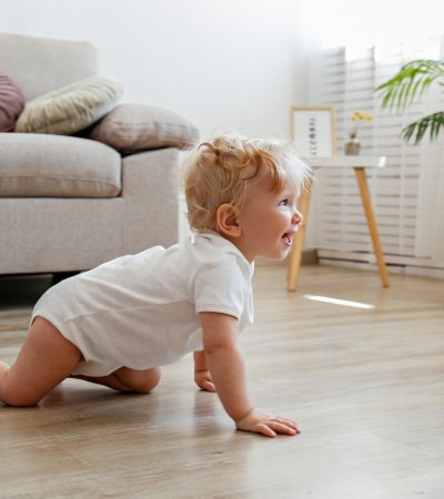 One year old child learning to crawl on the wooden floor of living room. Interior background. Adorable blonde little girl in pink crawlers at home. Close up, copy space for text.